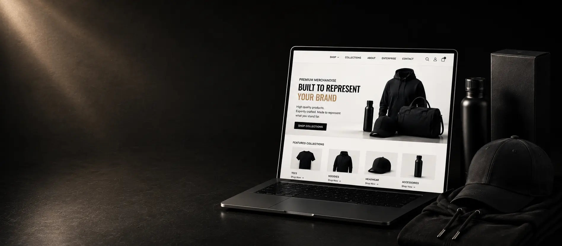 A laptop on a dark tabletop displaying a custom e-commerce website template for a clothing brand, surrounded by dark apparel and accessories under dramatic spotlighting.