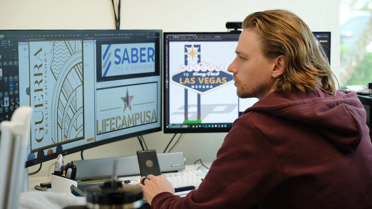 A man with blond hair sitting while designing several logos on two monitors.