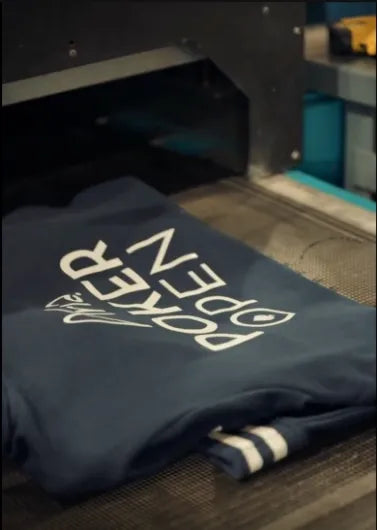 A navy blue sweatshirt with white 'POKER OPEN' text and a small logo, shown emerging from a black printing machine on a conveyor belt.