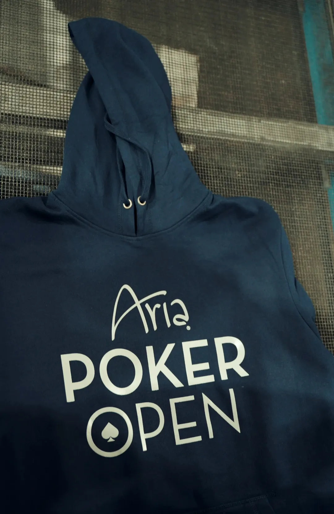 A navy blue hoodie featuring a white graphic print that reads 'Aria POKER OPEN' with a spade symbol, laid flat against a dark mesh screen background.