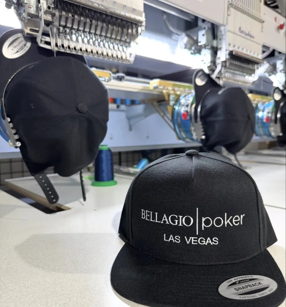 A black flat-brim baseball cap with white embroidered text reading 'BELLAGIO | poker LAS VEGAS' is shown in a textile or embroidery workshop setting, with sewing machines and other caps in the background.