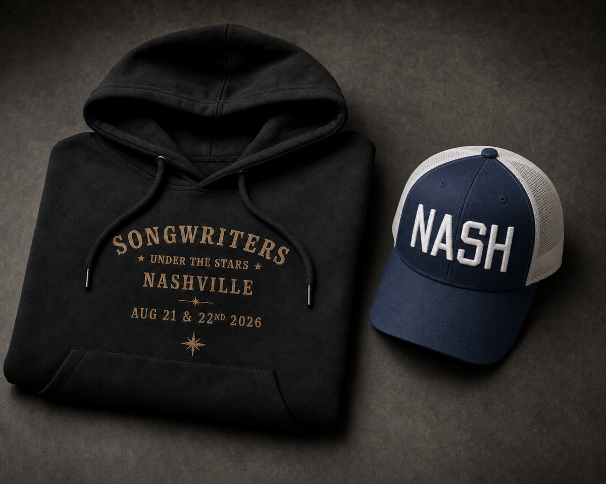 Songwriters Under The Stars tribute collection flat lay: folded black hoodie printed with gold Songwriters Under The Stars Nashville Aug 21 & 22nd 2026 text, next to a navy and white two-tone trucker cap embroidered NASH in white block letters