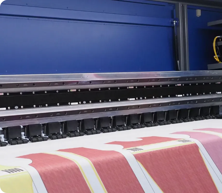 A large industrial textile printing machine with a blue upper housing and metallic guide rails, processing a white fabric sheet with pink and yellow patterned sections.