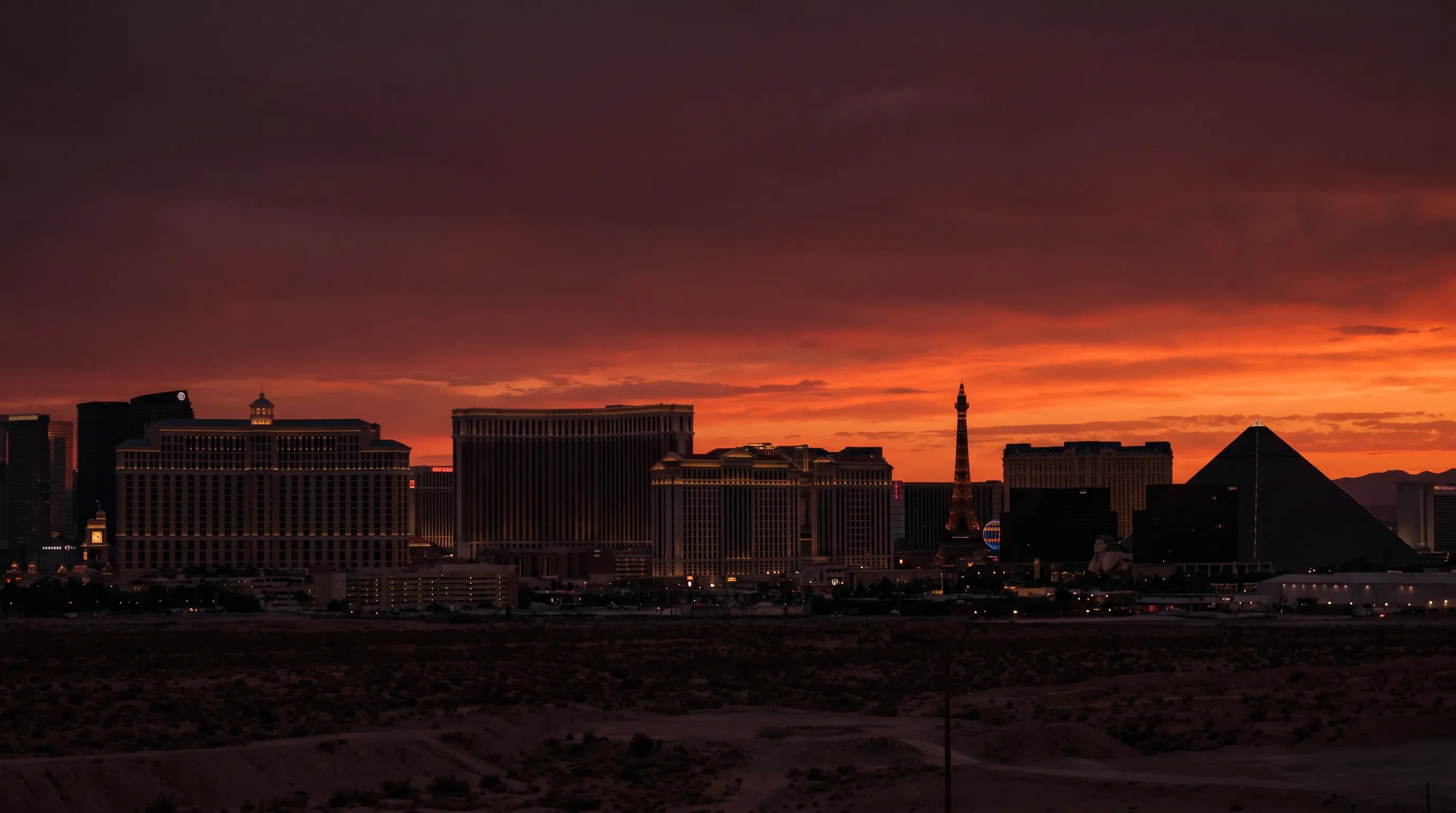 A dramatic sunset skyline of Las Vegas with silhouetted casino buildings, including the Eiffel Tower and pyramid, against a fiery orange and red sky.