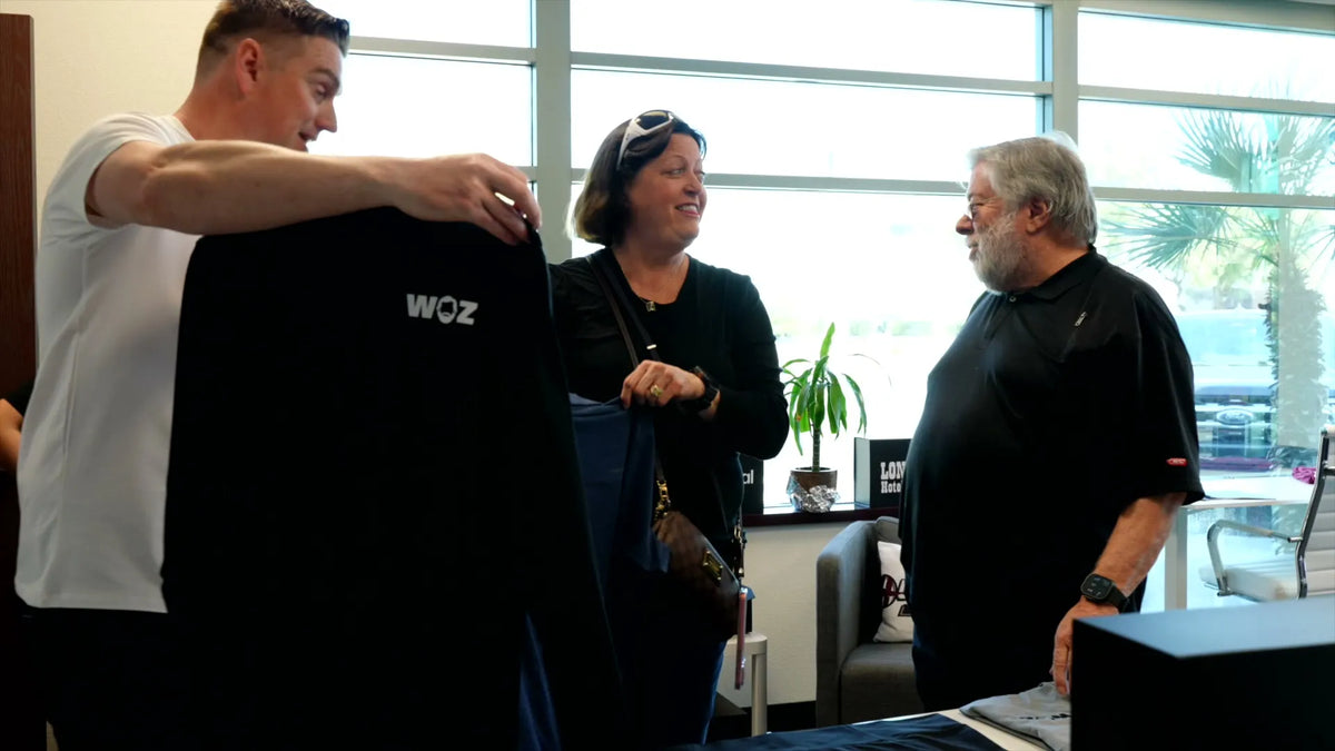 A man holds up a black t-shirt with 'WOZ' printed on the chest in an indoor setting, with two other men and a woman observing. Large windows and a potted plant are visible in the background.